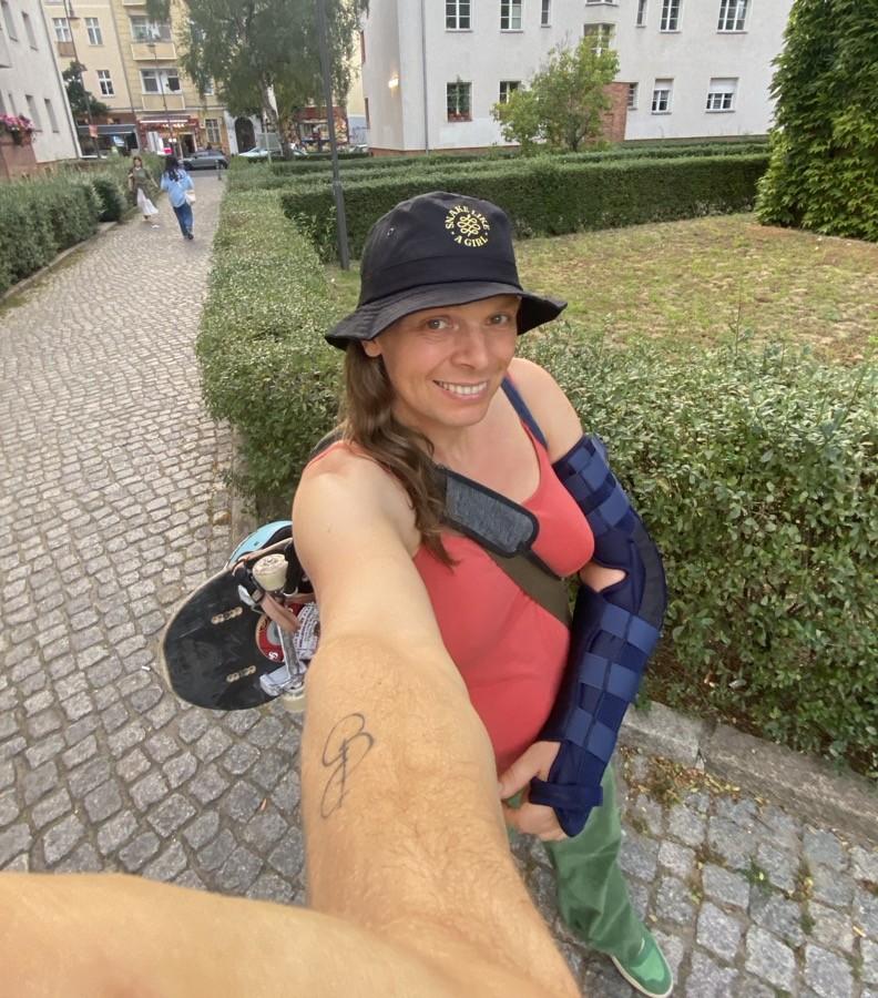 Me - a white trans-femme - walking outdoors and taking my streetboard with me. I'm smiling. The image shows my injured arm as well